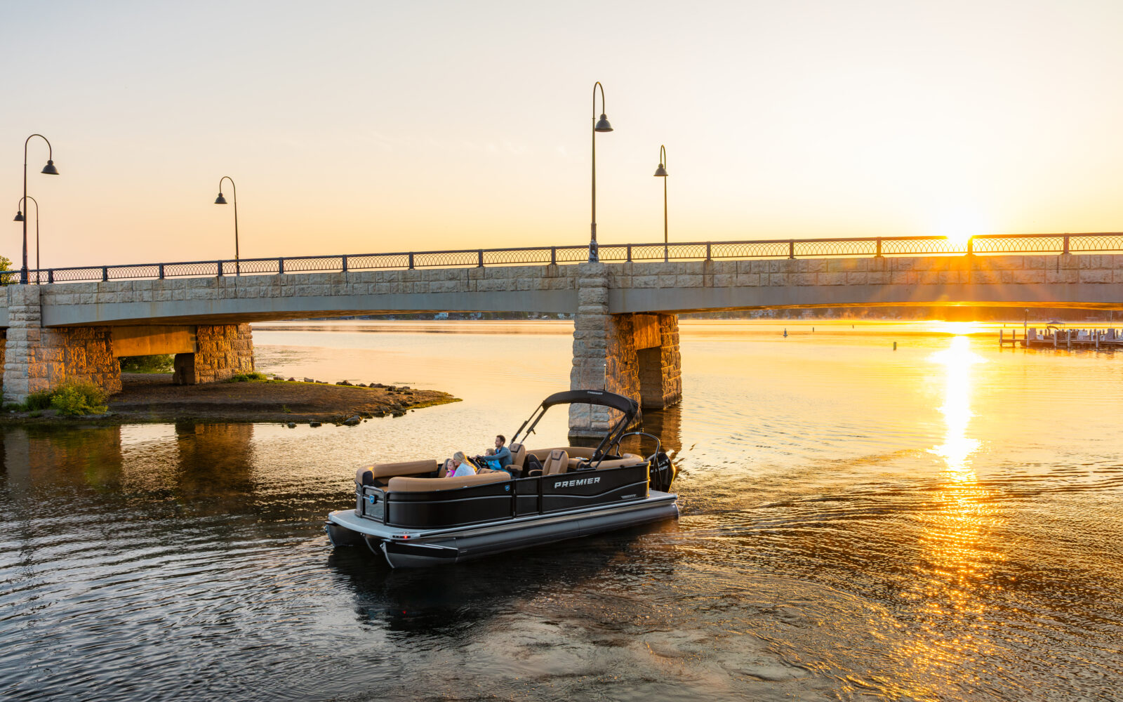 Premier Sunscape pontoon boat cruising beneath a bridge at sunset.