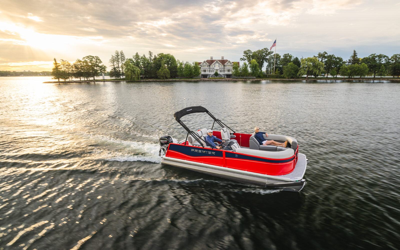 Red Premier Sunscape pontoon boat cruising across a lake at sunset.